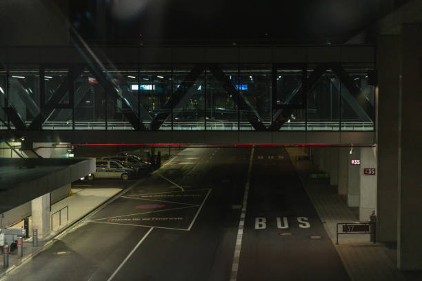 A dimly lit airport roadway at night featuring a modern pedestrian bridge with steel framework and glass walls. The scene includes an empty bus lane, parked police vehicles, and illuminated signs, creating a calm yet slightly mysterious nighttime airport atmosphere. The contrast between the artificial lights and dark surroundings adds a sense of travel, security, and urban infrastructure.