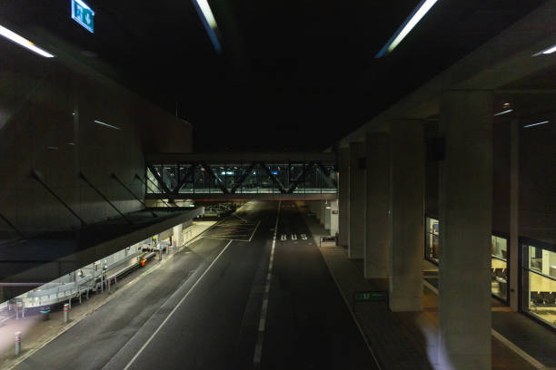 A dimly lit airport roadway at night featuring a modern pedestrian bridge with steel framework and glass walls. The scene includes an empty bus lane, parked police vehicles, and illuminated signs, creating a calm yet slightly mysterious nighttime airport atmosphere. The contrast between the artificial lights and dark surroundings adds a sense of travel, security, and urban infrastructure.