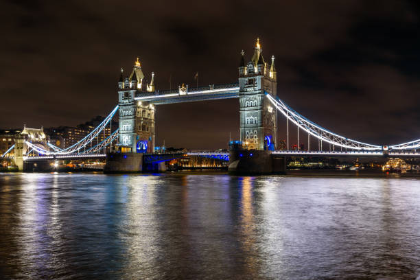 London, England, United Kingdom  - December 17, 2022: Profile shot of the beautifully illuminated Tower Bridge over the Thames River at night with city lights in distance.