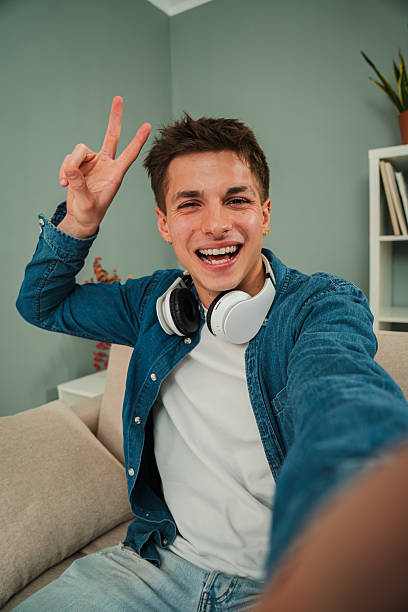 istock Vertical Young adult man smiling taking a selfie at home. Friendly guy shooting a photo looking at camera with perfect white teeth smile at living room. Individual portrait of a real caucasian male 2204765607