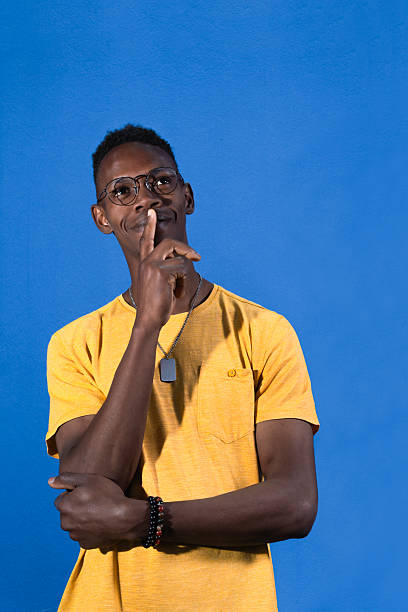 Pensive young african man making a silence gesture with his finger on his lips, looking up on a blue background