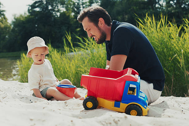istock Boy with a hat plays in the sandbox on the beach, pouring sand into a bucket, while dad supervises, creating outdoor memories. 2204774030