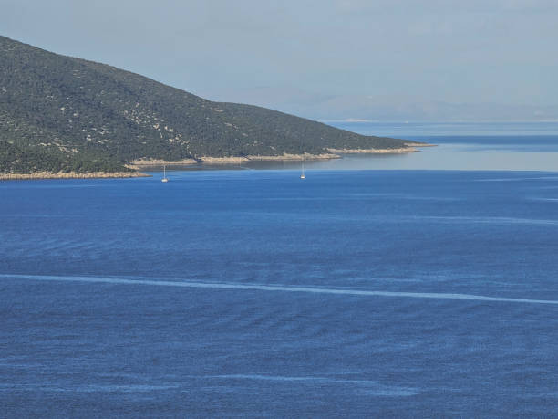 A peaceful seascape featuring two sailboats floating on the calm, deep blue waters near a lush green peninsula. The coastline gently curves into the distance, meeting the soft horizon where the sea blends with the sky. The image captures the essence of serenity and maritime adventure, perfect for those who love the ocean’s vast beauty.