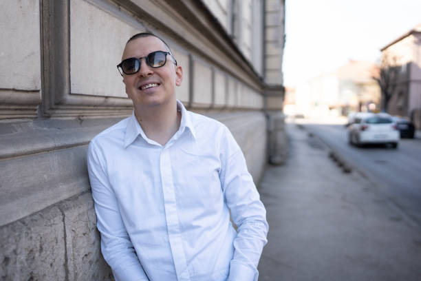 A cheerful man leans against a building in the sunlight, wearing a white shirt and sunglasses. The background shows a street scene with vehicles and urban architecture, suggesting a casual outdoor setting.