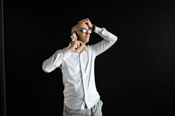 A man in glasses and a white shirt holding a phone and touching his head under apparent stress or frustration, standing against a dark background. Illustrates emotions of concern and unease.