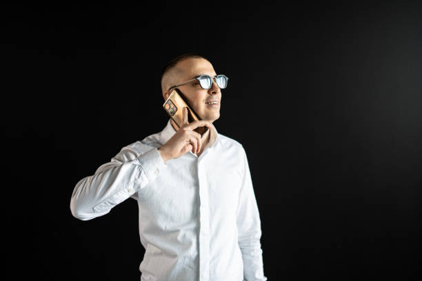 A business professional wearing a white shirt and glasses engages in a phone call, exuding confidence in a minimal dark backdrop setting. Ideal for business, communication, and modern professional themes.