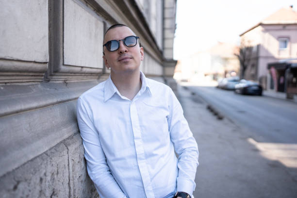 A young man wearing sunglasses and a white shirt leans against a wall in an urban environment, exuding confidence and relaxation in the spring sunlight.
