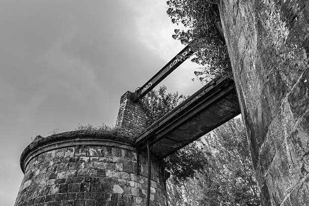 An old rusty metal bridge sprayed with graffiti connecting two abandoned stone brick towers with overgrown foliage shot in black and white against a clear sky background.