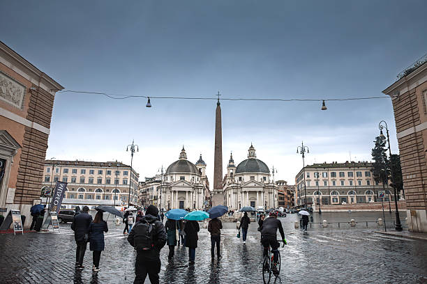 Rome, Italy - January 18, 2025: Vibrant street scene from Piazza del Popolo in Rome, Italy, showing pedestrians using umbrellas on a rainy day. Central to the image is the Flaminio Obelisk, surrounded by the piazza's classic symmetrical Italian architecture. This photograph beautifully captures urban life, Roman heritage, and the tourist atmosphere of this historic landmark under wet conditions.