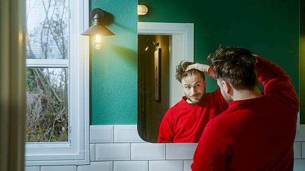 istock Young man in a red sweater looking at himself in the mirror with a concerned expression, touching his hair as he contemplates hair loss, aging, or self-image issues 2204809485
