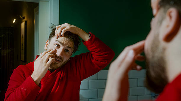 istock Young man in a red sweater examining his new eyebrow piercing in the mirror with a concerned expression, feeling discomfort or uncertainty about the fresh piercing 2204809722