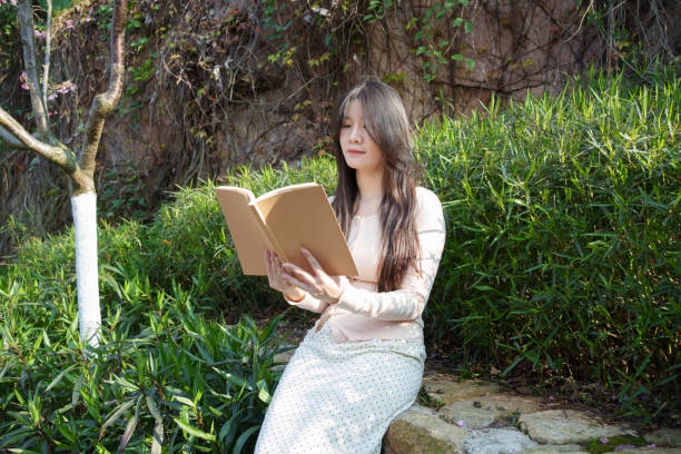 A beautiful woman in her twenties is sitting in nature, reading a book. She is enjoying a lovely sunny day alone, feeling content and smiling. The view from the top of the cliff overlooks the beautiful landscape.