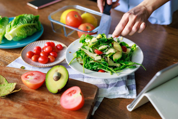 A person arranges cherry tomato fresh and avocado slices on a bed of arugula and mixed greens while using a tablet for guidance. Surrounded by cherry tomatoes, lemons, and lettuce, this vibrant salad highlights fresh and healthy eating.