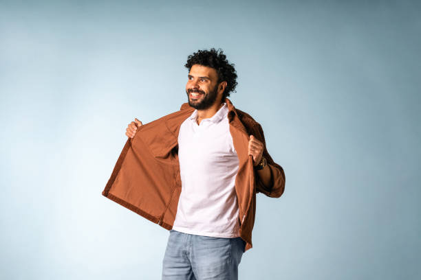 A mid-adult Middle Eastern male smiling confidently while wearing a casual brown jacket and white shirt, standing against a light blue background in bright studio lighting.