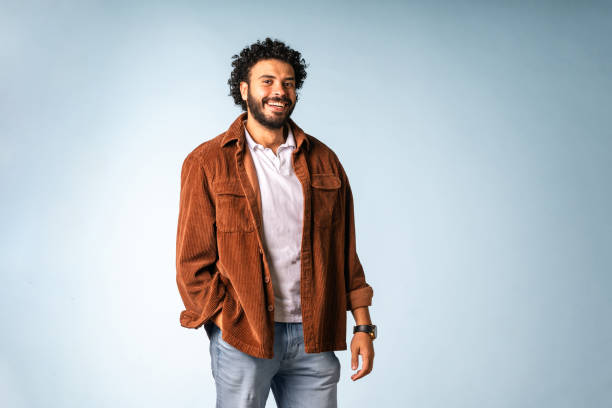 A cheerful Middle Eastern man in his 30s wearing a brown jacket, pale shirt, and jeans poses confidently against a light blue background.