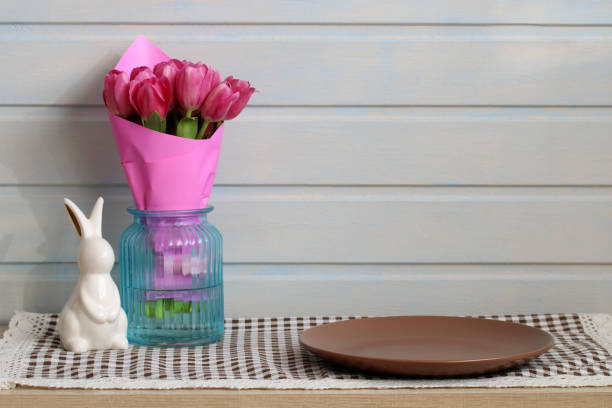 Pink tulips bouquet in blue glass vase on checkered tablecloth with empty brown plate and decorative rabbit figurine. Easter still life