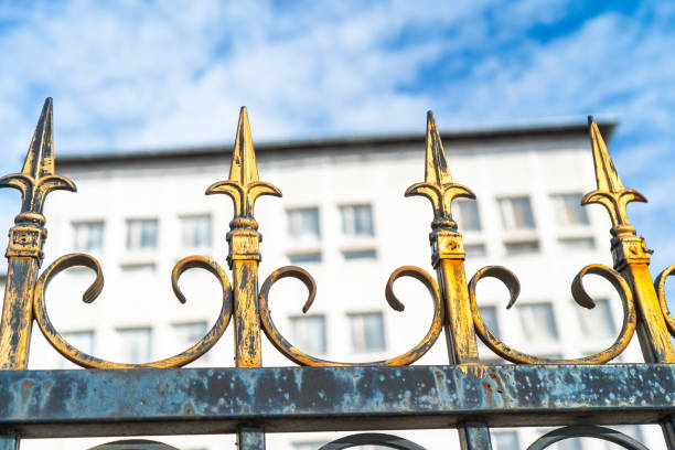 An ornate metal fence with golden spear-shaped tips and intricate scrollwork stands in the foreground, showing signs of weathering and rust. Behind it, a white multi-story building with rows of windows is slightly blurred, contrasting the decorative yet aged barrier. The blue sky adds depth to the composition, highlighting the architectural contrast between the fence’s craftsmanship and the modern structure beyond.