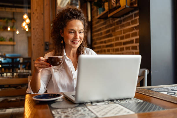 istock Smiling woman working on laptop and drinking coffee in cafe 2204839908