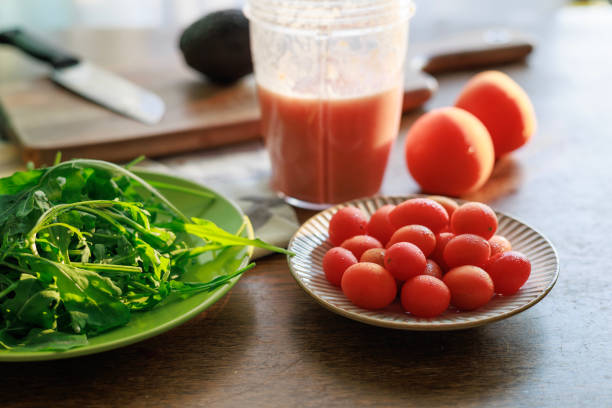 A refreshing homemade tomato smoothie in a blender cup, surrounded by fresh ingredients including cherry tomatoes, leafy greens, and whole tomatoes on a wooden table. Natural light enhances the vibrant colors, highlighting the concept of healthy eating and fresh, organic food preparation.