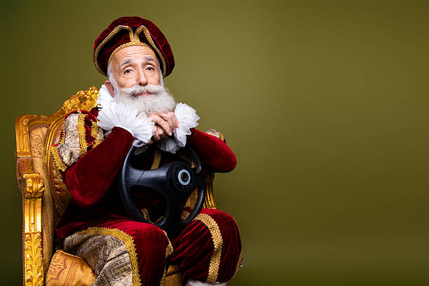 istock Senior man dressed as medieval aristocrat holding steering wheel with khaki background, blending historic costume with modern concept 2204854549