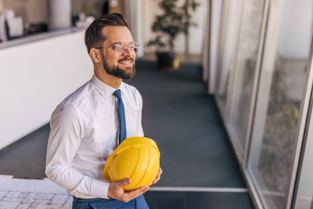 istock Smiling architect holding hardhat looking out large windows in office 2204869718