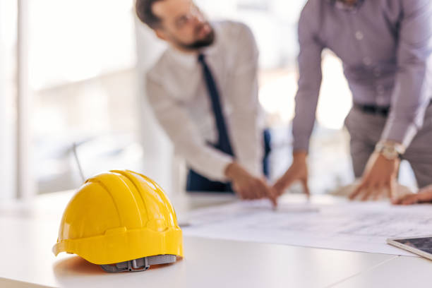 istock Yellow hard hat sits on desk while architects discuss blueprint 2204869861