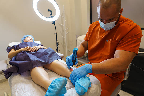 Doctor using laser device to remove tattoo from leg of young woman lying on medical couch in beauty salon
