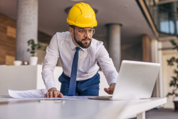 istock Architect wearing hardhat working on laptop and blueprints in office 2204872444