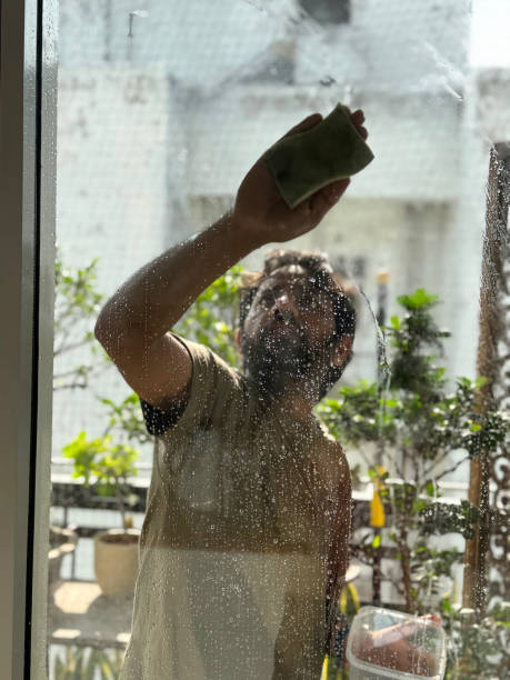 istock Image of Indian man on high residential balcony, window cleaner using sponge and soapy water to clean glass window pane, safety railing, apartment block background, focus on foreground 2204884044