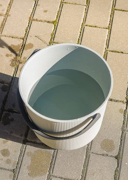 istock Gray Plastic Bucket Filled with Water on Paved Stone Surface, Bright Sunlight 2204889437