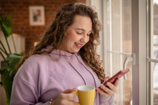 istock Plus size young woman enjoying a cozy moment at home while sipping coffee and checking her phone 2204896377