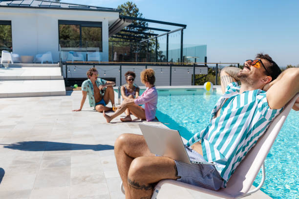 A group of friends enjoying a sunny day by the pool, talking and connecting. A man with a laptop exemplifies a casual working lifestyle.