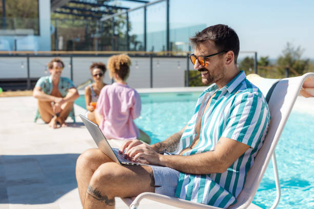 A man sits by a pool using a laptop, with friends relaxing nearby. This image captures the essence of modern freedom, a creative and relaxed working atmosphere outdoors.