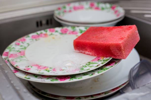 Hand with pink sponge washing floral-patterned dishes in metal sink. Tiled kitchen background with hanging utensils. Everyday household chore captured in action