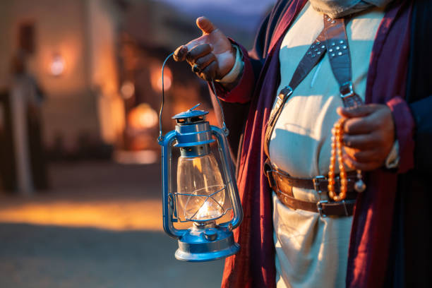 A traditional Saudi Arabian lantern glows in the night. A man dressed in traditional attire holds an old-fashioned lantern, illuminating the night with a warm glow. On his other hand, he holds prayer beads, symbolizing spirituality and heritage. The background features a softly lit, historic village setting, evoking a sense of nostalgia and cultural richness in Saudi Arabia. High quality photo