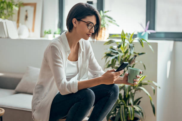 Pretty woman using smartphone while drinking coffee sitting on stool at home