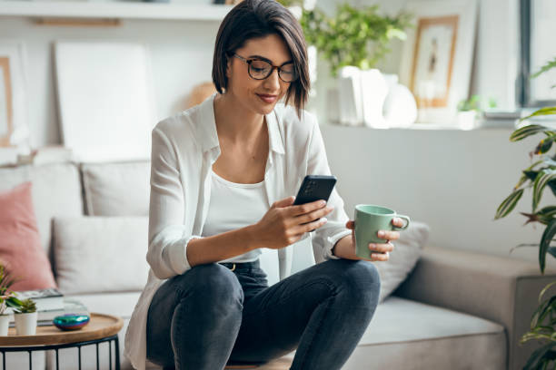 istock Pretty woman using smartphone while drinking coffee sitting on stool at home 2204912212