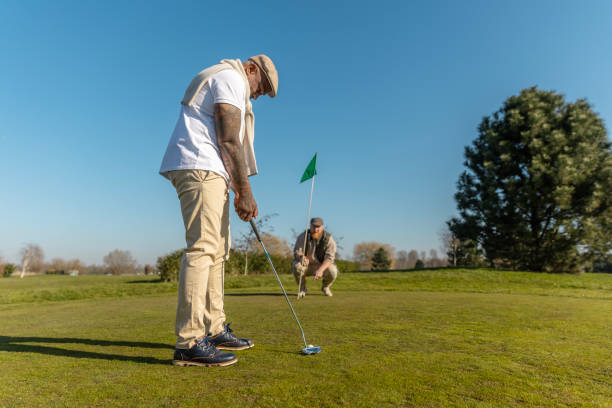 Asian male golfer is putting on the green while an elderly golfer stands nearby cheering and watching, capturing a moment of support and excitement on a scenic golf course. Golf course in morning.