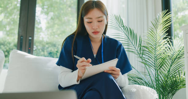 istock Young Asian female nurse in blue scrubs is deeply focused on her laptop while taking notes during an online medical course in living room at home. Nurse sustainable lifestyle concept. 2204921937