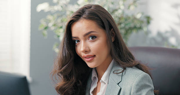 The efficient and beautiful secretary sits at her desk, writing emails to clients, tapping her fingers on the keyboard, and smiling at the camera with a professional yet friendly demeanor.
