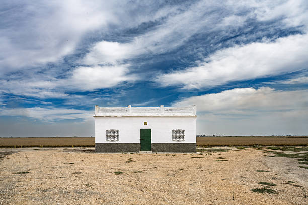 A rural white house located amidst vast fields under a clear blue sky.
