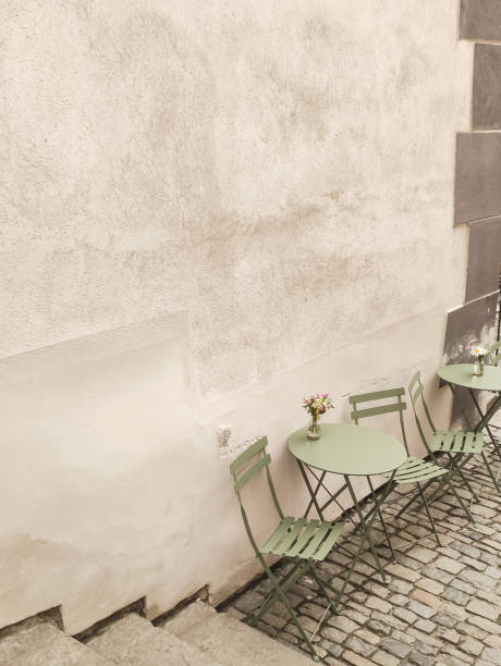 Green bistro tables and chairs sit empty on a cobblestone street alongside a weathered wall in cesky krumlov, czechia, waiting for customers to arrive
