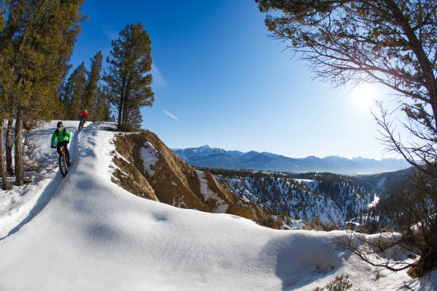 A man leads a woman down a hill during a winter fat bike ride along the edge of a river canyon on a sunny morning near Invermere, British Columbia, Canada. They both wear cycling helmets, warm winter clothing, and backpacks. Fat bikes are mountain bikes with large wheels and tires for riding on the snow.