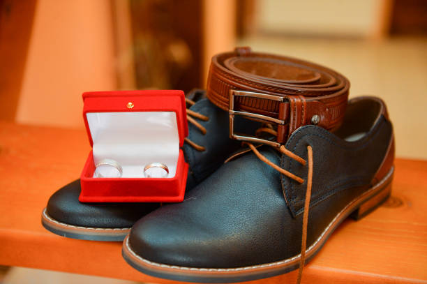 two white gold wedding rings in a red festive and elegant box that is on the groom's shoes and next to it is a brown leather belt