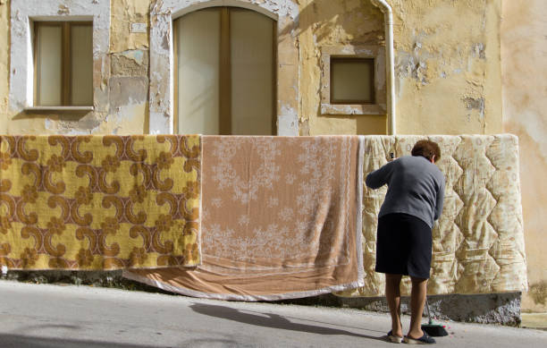 Sortino, Sicily, Italy: A senior woman sweeps the street in downtown Sortino as she airs out her blankets on her balcony. The village is in Siracusa Province.