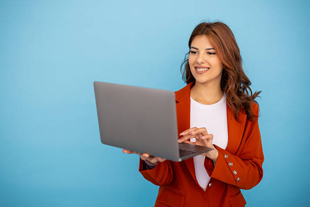 istock Young businesswoman using laptop, smiling and looking at screen on blue background 2204958240