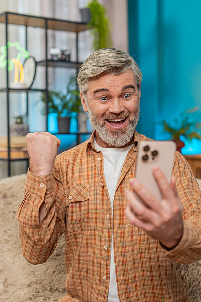 istock Mature man celebrating work success and online game victory while using smartphone on sofa at home 2204976189