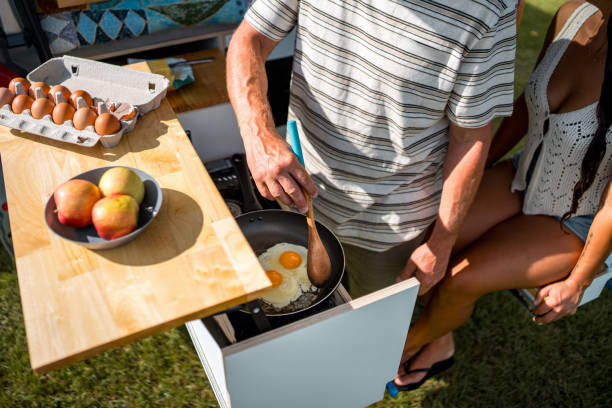 Caucasian father cooking breakfast outdoors on a sunny camping trip, frying eggs beside fresh fruit and ingredients on a wooden counter.
