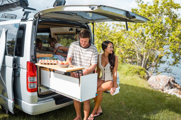 A Caucasian father prepares breakfast outdoors near a campervan during a family camping trip. A woman relaxes nearby, surrounded by lush greenery and a scenic lake view.