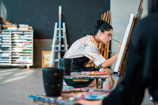 Side-profile portrait of a young Middle Eastern woman in a white shirt concentrating on painting at an easel, surrounded by colorful art supplies in a modern studio space.